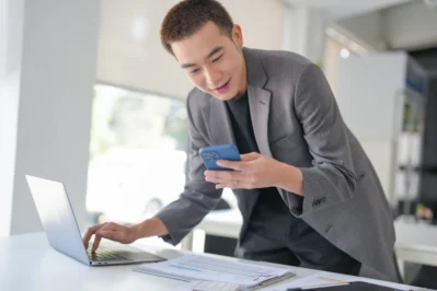 Homem sorrindo usando o celular e laptop em mesa com documentos, ilustrando facilidade do horário Pix para pagamentos.