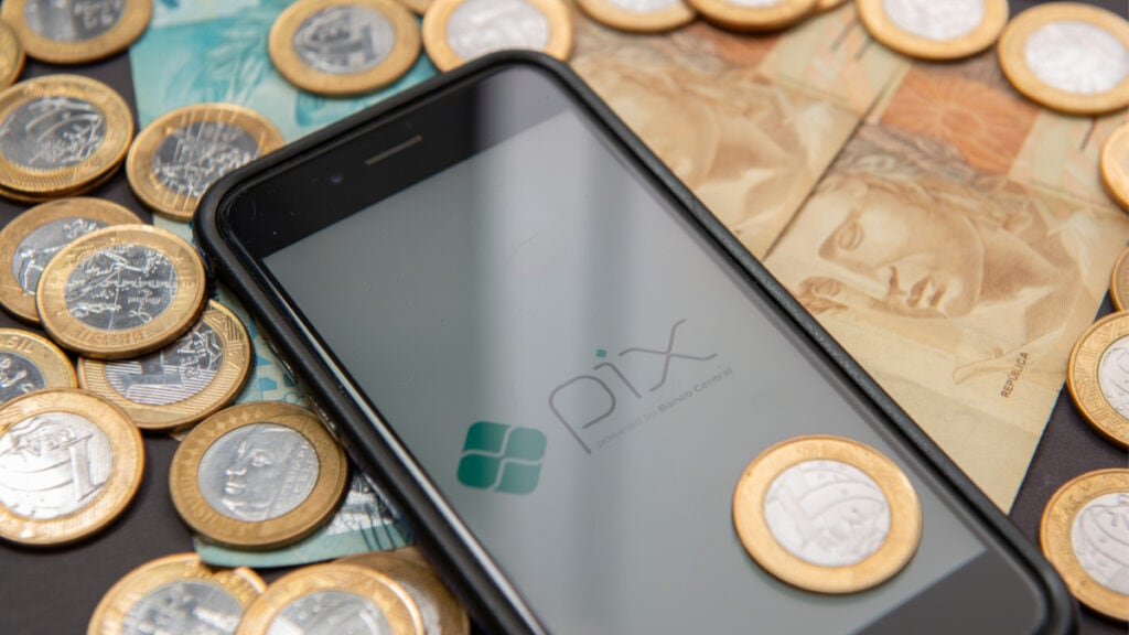 Celular sobre uma mesa exibindo na tela o logotipo do Pix. Ao seu redor, há algumas cédulas de dinheiro e algumas moedas de 1 real, representando o Pix Troco.