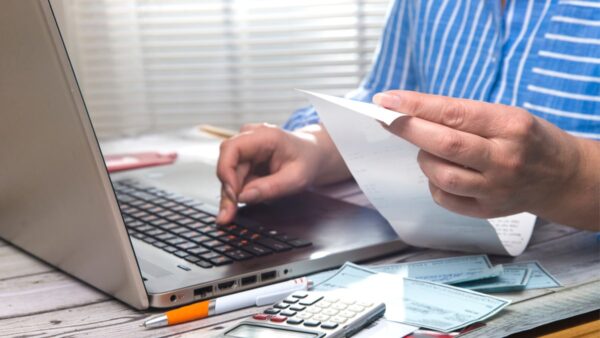 Pessoa segura uma conta entre os dedos da mão esquerda, enquanto digita no teclado do notebook com os dedos da mão direita. Ao seu redor, há alguns papéis, uma calculadora e uma caneta. O objetivo é representar o controle de contas a pagar.