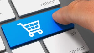 Teclado de computador, com foco em um botão azul com o símbolo de carrinho de compras sendo acionado por um dedo humano. A ideia é representar visualmente o conceito de one click buy (compra com um clique).