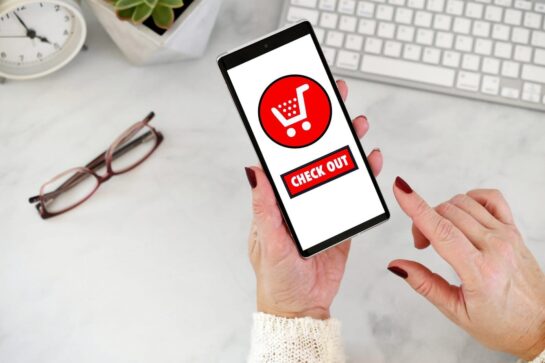 Na imagem tem mão segurando um celular, na tela está escrito chek out e tem um carrinho desenhado. Ela está finalizando uma compra no checkout transparente de uma loja virtual.