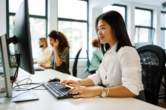 Funcionária sentada em frente ao computador, digitando, sorrindo e utilizando ERP SaaS para gerenciar o negócio.