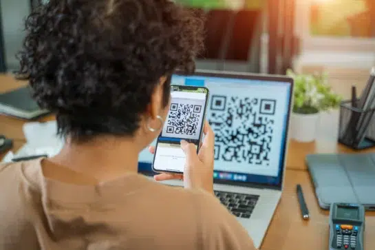 Mulher escaneia um QR code na tela do celular, gerado no notebook, indicando o uso de um gerador de QR code Pix.