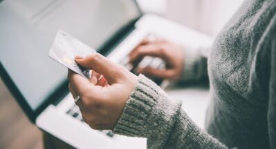 Na imagem uma mulher branca vestindo uma blusa de lã cinza comprida, está trabalhando em seu computador. A mulher, está com um cartão de crédito em uma das suas mãos, isso significa que ela pode aceitar cartão de crédito como forma de pagamento em seu negócio ou mesmo, pode estar efetuando uma compra online.