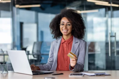 Uma empreendedora está em seu escritório, mexendo no notebook e segurando um cartão. Ela está pesquisando o que é cartão de crédito corporativo e quais benefícios ele possui para sua empresa.
