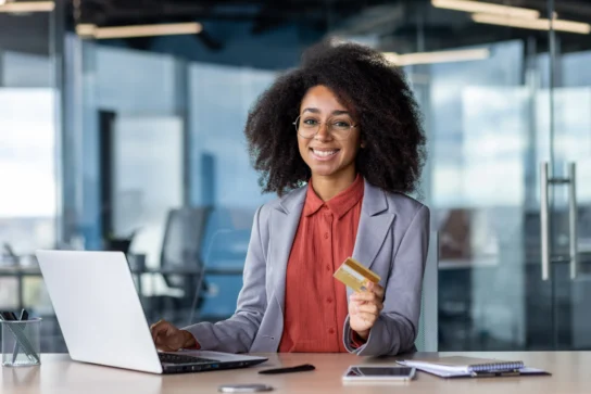 Uma empreendedora está em seu escritório, mexendo no notebook e segurando um cartão. Ela está pesquisando o que é cartão de crédito corporativo e quais benefícios ele possui para sua empresa.