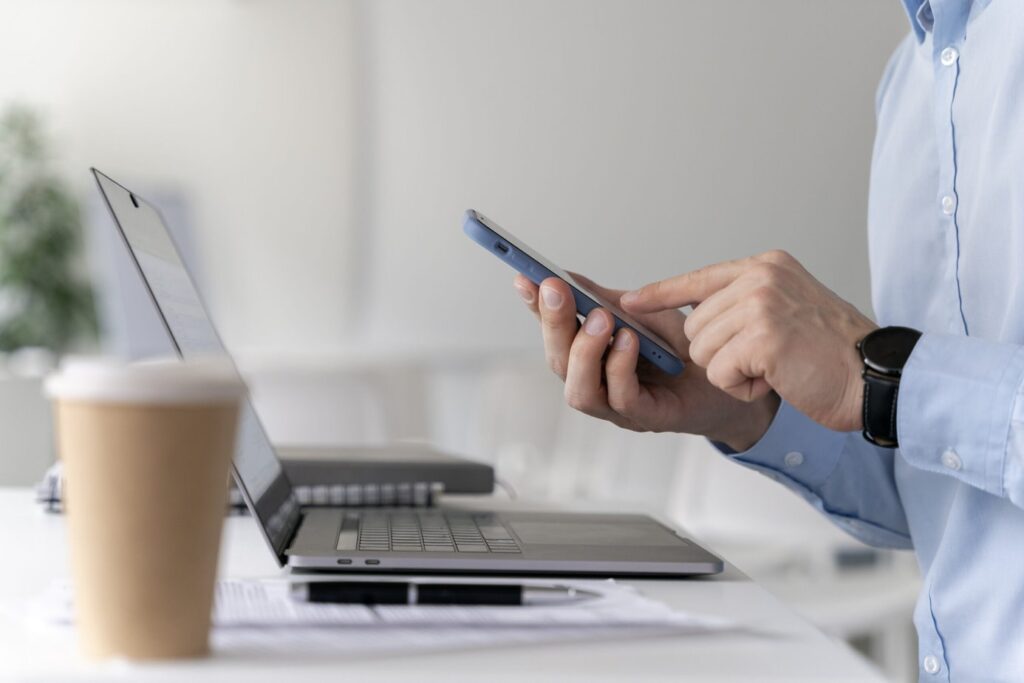 A imagem mostra as mãos de um homem segurando o celular, ele está sentado em frente ao notebook. Ilustrando a melhor plataforma de antecipação de recebíveis.