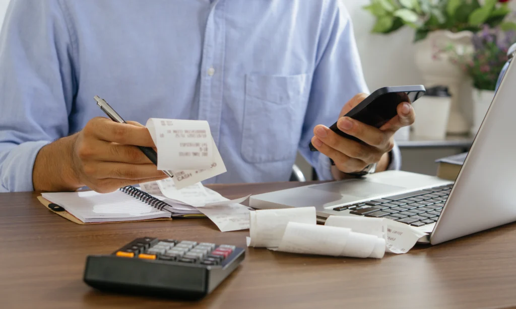 Homem com contas na mão, celular e calculadora na mesa. Ele está checando mensagens recebidas pelo sistema de cobrança via WhatsApp.