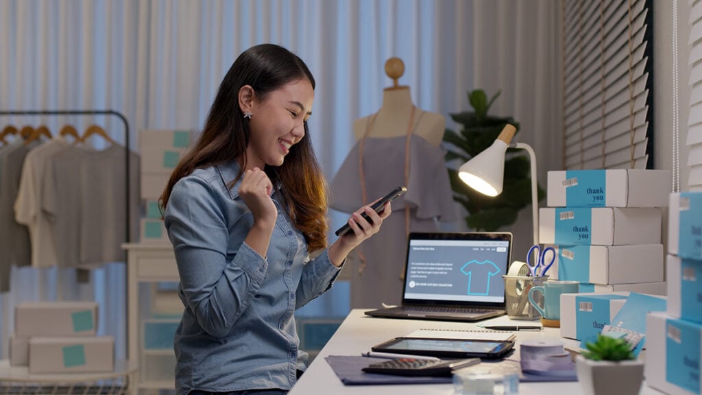 Na imagem, uma mulher feliz, com o notebook sobre a mesa e o celular na mão, fazendo venda por link de pagamento.