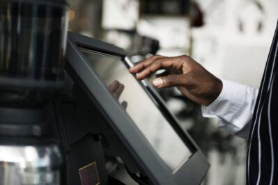 Homem está em pé atrás do caixa, ele está apontando para a tela do computador, onde manuseia o sistema de automação comercial.