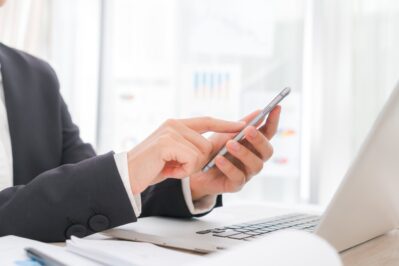 Na imagem, temos as mãos de um homem sobre a mesa de seu escritório, segurando um celular em uma das mãos e com a outra está manuseando o notebook. Ele está procurando como gerar link de pagamento.