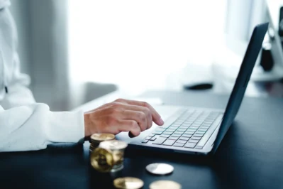 Homem utilizando uma plataforma de cobrança recorrente no notebook. Ao lado há moedas de ouro em cima da mesa.