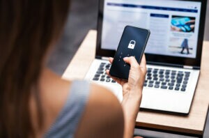 A imagem mostra uma mulher com o celular na mão, em frente ao computador, fazendo a autenticação multifator para entrar no seu app.