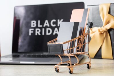 Na imagem, temos um notebook em cima de uma mesa, com a escrita "Black Friday" na tela. Ao lado, há uma caixa de presente preta. Mais à frente, temos um carrinho de compras pequeno com alguns cartões de crédito dentro. Isso representa as formas de pagamento para Black Friday.