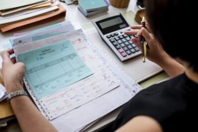 Na imagem, temos uma empreendedora com seus boletos em cima da mesa, com uma calculadora na mão. Ela está entendendo como gerar boleto parcelado, pra ver se vale a pena para o seu negócio.