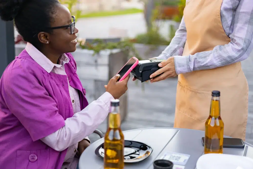Cliente realiza pagamento via Pix garantido em maquininha, utilizando celular em mesa de bar com bebidas.