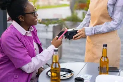 Cliente realiza pagamento via Pix garantido em maquininha, utilizando celular em mesa de bar com bebidas.