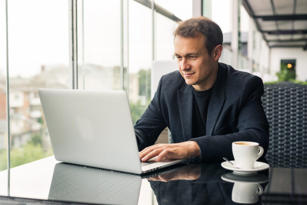 A imagem mostra um empresário em sua mesa do escritório, mexendo no computador. Ao seu lado há uma xícara de café. Ele está pesquisando uma conta digital para empresa ME.