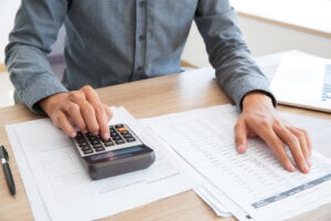 A imagem mostra um empresário em sua mesa no escritório. Ele está analisando o registro do fluxo de caixa, utilizando uma calculadora para calcular seu imposto de renda de pessoa jurídica.