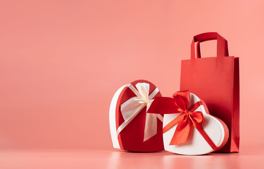 Na imagem tem um fundo rosa, e nela tem dois pacotes de presente com formato de coração e uma sacola vermelha, representando como vender mais no dia dos namorados.