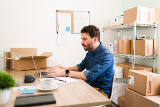 A imagem mostra um empreendedor na mesa do seu escritório. Atrás dele, há vários produtos em caixas para envio. Ele está mexendo em um notebook, pesquisando como vender mais no dia dos pais.