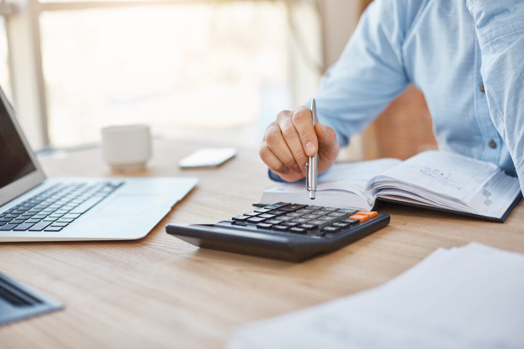 A imagem mostra um empreendedor em seu escritório. Em sua frente está o notebook, um caderno e uma calculadora ao lado. Ele está pesquisas e cálculos para entender como pagar menos impostos.