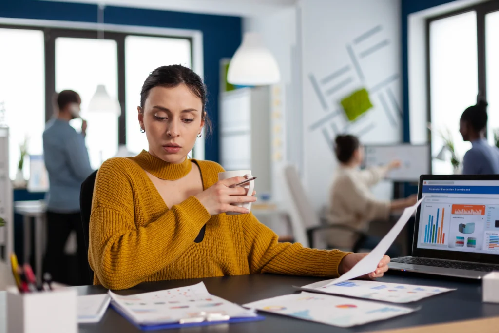 Empreendedora sentada à mesa, segurando xícara enquanto avalia gráficos. Está realizando a gestão financeira para pequenas empresas.