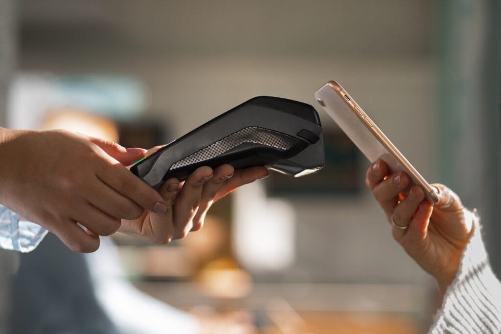 A imagem mostra uma atendente finalizando uma compra. Ela está com a maquininha de cartão e o seu consumidor está com o celular próximo à tela da máquina, esperando o processo de pagamento ser concluído.