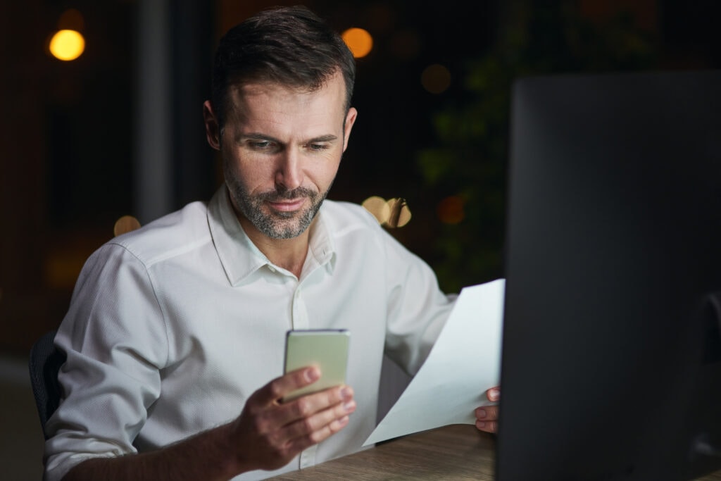 A imagem mostra um empreendedor no seu escritório, em frente ao computador, segurando uma fatura e um celular. Ele esta testando sua nova conta digital para emissão de boletos.