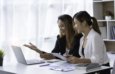 Gestoras analisando funções da gestão financeira em um laptop, com gráficos e cadernos em uma mesa iluminada por luz natural.