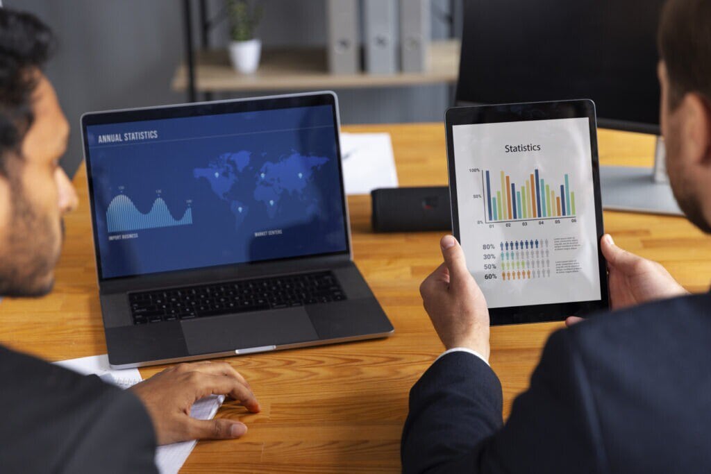 A imagem mostra dois homens, um segura um tablet, o outro está em frente ao notebook. Eles estão vendo os gráficos de resultados do manual de gestão financeira.