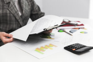 Na imagem, temos um homem com um livro na mão, em sua frente em uma mesa cheia de papel e uma calculadora. Isso mostra o que é gestão financeira de um negócio.
