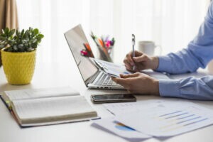 A imagem mostra um empreendedor em seu escritório, com um computador e diversos gráficos impressos sobre a mesa. Ele está entendendo qual é a função da gestão financeira.