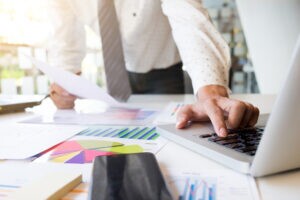 Homem em frente a uma mesa cheia de papéis com gráficos e um notebook, representando como fazer a gestão financeira de uma empresa.