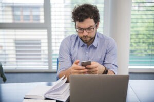 Empreendedor em frente ao computador. Ele está mexendo em seu celular, buscando por um aplicativo de gestão financeira.
