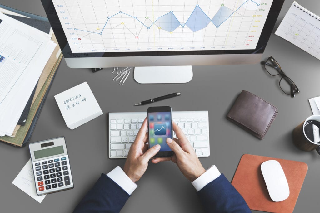 Empreendedor em sua mesa do escritório, com seu computador ligado e um celular na mão. Ele está utilizando ferramentas de gestão financeira.
