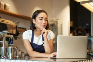 Uma empreendedora está no balcão, mexendo em seu computador. Ela está pesquisando sobre gestão financeira para restaurantes.