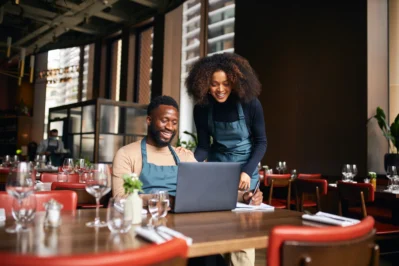Duas pessoas com aventais analisando dados financeiros em um laptop enquanto cuidam da gestão financeira para restaurantes.