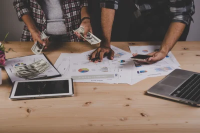 Duas pessoas lidando com dinheiro, gráficos e tecnologia em mesa de trabalho, ilustrando a gestão financeira para startups.