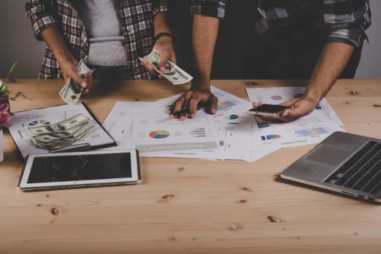 Duas pessoas lidando com dinheiro, gráficos e tecnologia em mesa de trabalho, ilustrando a gestão financeira para startups.