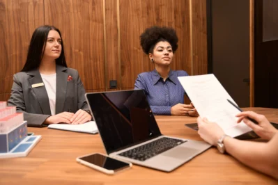 Mulheres trabalhadores em reunião. Em frente delas tem outra pessoa, fora de cena, com um computador em cima da mesa. Essa terceira pessoa ensina como consultar o CNPJ de uma empresa, com documentação no papel.