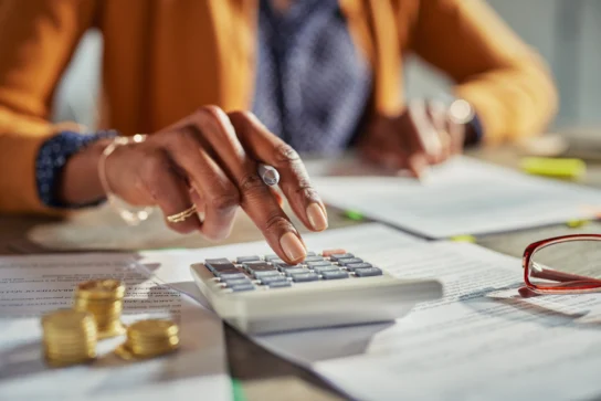 Mão usando calculadora em ambiente de trabalho com documentos e moedas sobre a mesa, representando como funciona IRPJ.