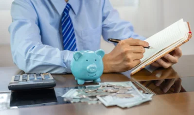 Homem anotando receitas e despesas da empresa em caderno, com porquinho azul, moedas, cédulas e calculadora sobre a mesa.