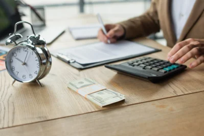 Homem calculando finanças com relógio e dinheiro sobre a mesa, representando o tempo de compensação de boleto.