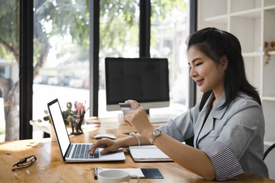 Funcionária de empresa está utilizando o cartão de crédito para CNPJ para realizar compras. Está sentada em uma mesa de escritório, digitando no computador.