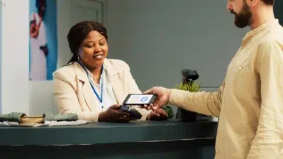 Homem realizando pagamento com celular em terminal, atendente sorrindo verificando se não houve pagamento duplicado