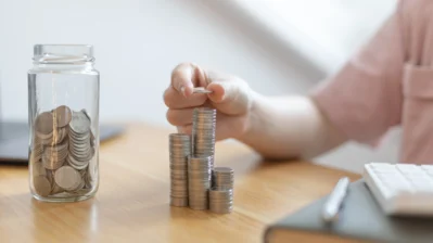 Mulher está aprendendo como economizar dinheiro, empilhando moedas em cima de uma mesa e guardando em um pote.