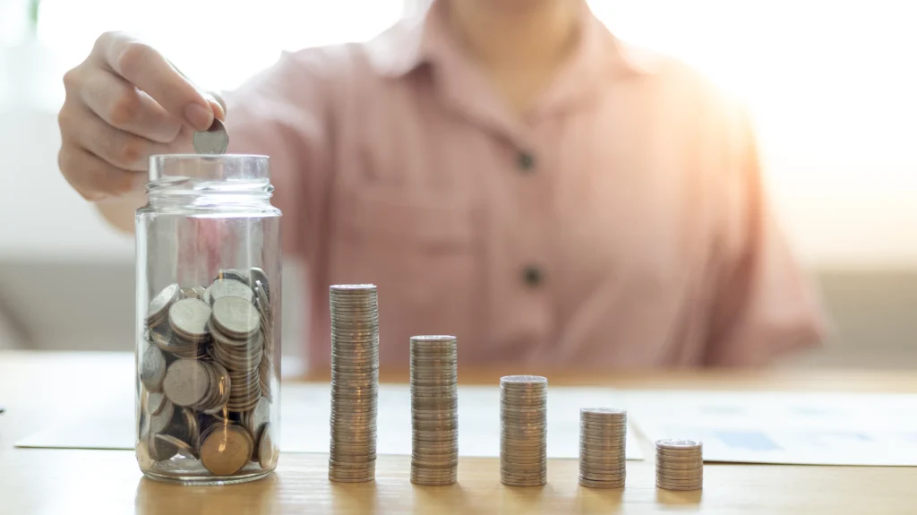 Pessoa colocando moedas em um pote ao lado de pilhas de moedas, exemplificando o consumo consciente de dinheiro.