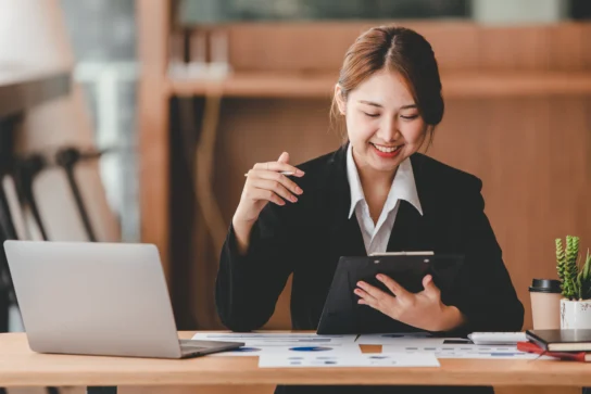 Empresária sorrindo, com caneta na mão e um notebook na mesa, analisando gráficos para escolher a melhor conta PJ sem anuidade.