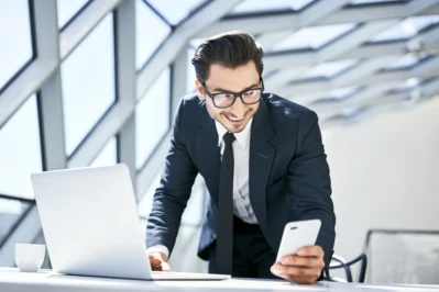 Empresário de óculos, terno e gravata, sorrindo para o celular e laptop, escolhendo a melhor conta PJ para sua empresa.